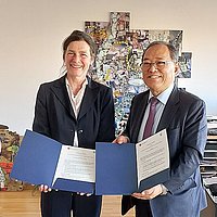 Auf dem Foto sind ASH-Präsidentin Bettina Völter und Shin-Il Yoon, Präsident der Kangnam University, zu sehen. Sie stehen nebeneinander im Büro der Präsidentin und halten gemeinsam den unterzeichneten Kooperationsvertrag in den Händen 